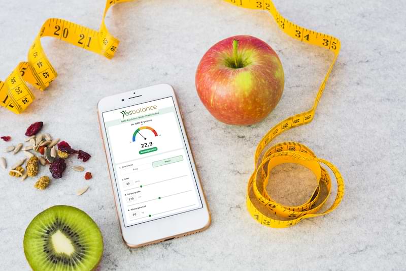 BMI Rechner von YesBalance auf einem Smartphone, daneben Apfel, Kiwi, Maßband und Trockenfrüchte